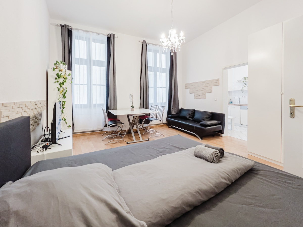 A spacious bedroom features a large bed with neatly arranged linens and a rolled towel atop. The room opens to a combined living and dining area, which includes a round dining table and modern seating. Large windows provide natural light, complemented by elegant curtains.