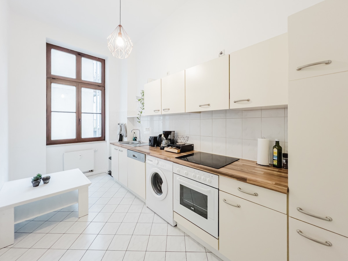 A well-equipped kitchen features white cabinetry and a wooden countertop. Essential appliances, including a stove, oven, dishwasher, and washing machine, are present. Natural light filters through a large window adorned with simple white curtains, illuminating the tiled flooring and a small table in the corner.