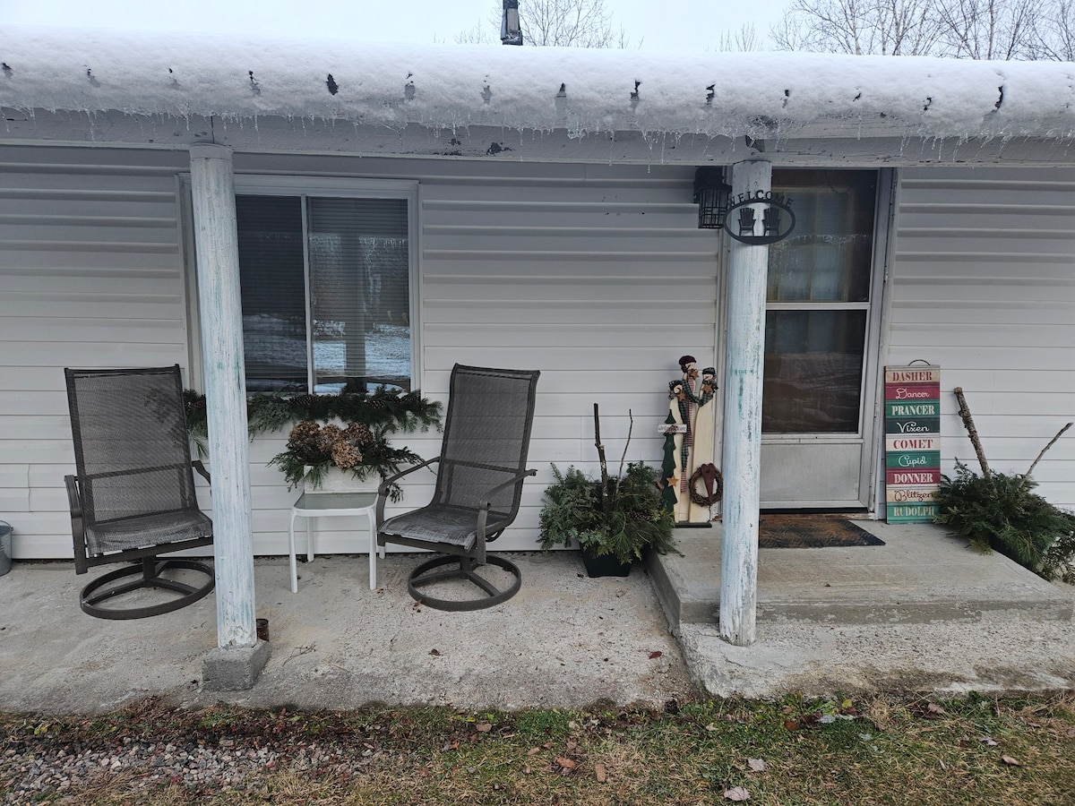 A welcoming entrance is seen, featuring two rocking chairs placed beside a small table, adorned with potted greenery. The door is framed by seasonal decorations and a sign indicating the unit number. Snow is gently visible on the ground, suggesting a winter ambiance.