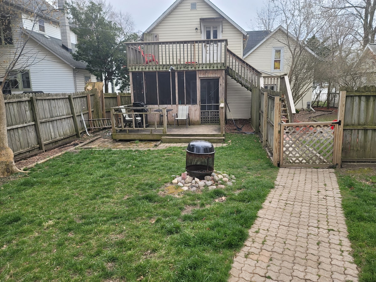 A fenced backyard features a grassy area, a stone fire pit surrounded by rocks, and a wooden deck attached to the house. The outdoor seating area with a table and chairs is positioned on the deck, offering a comfortable space for relaxation.