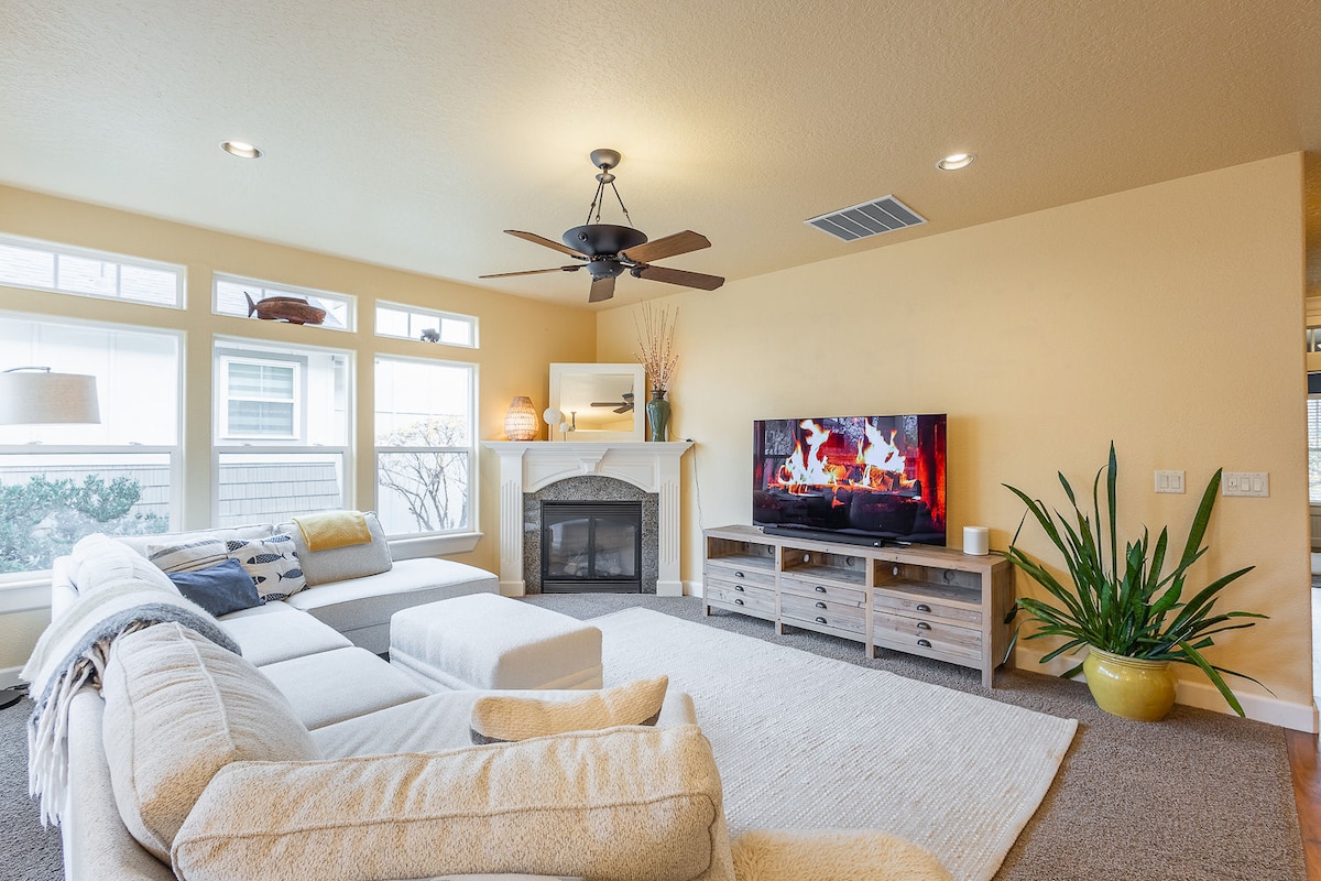 A light-filled living area features a spacious sectional sofa arranged around a central area rug. A large television displays a cozy fireplace scene. Natural light enters through multiple windows, complemented by a ceiling fan and a decorative houseplant in the corner.