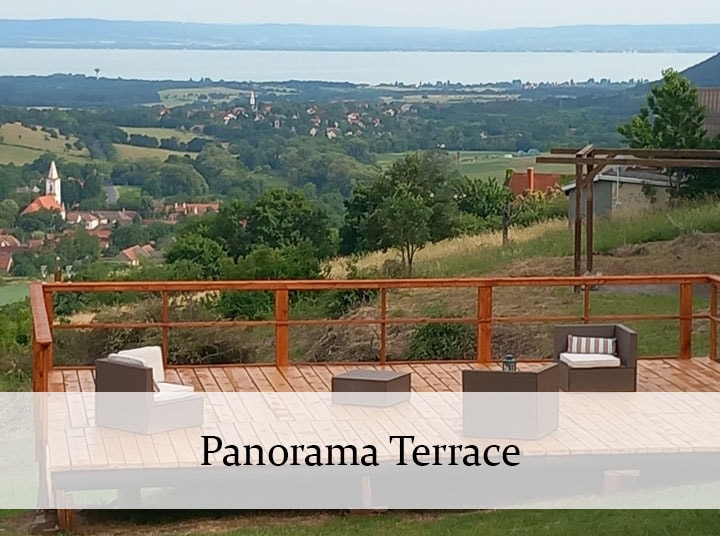 A spacious panorama terrace offers stunning views of Lake Balaton and the surrounding landscape. Outdoor lounge furniture is arranged on a wooden deck, providing an inviting space for relaxation and enjoyment of the natural beauty.