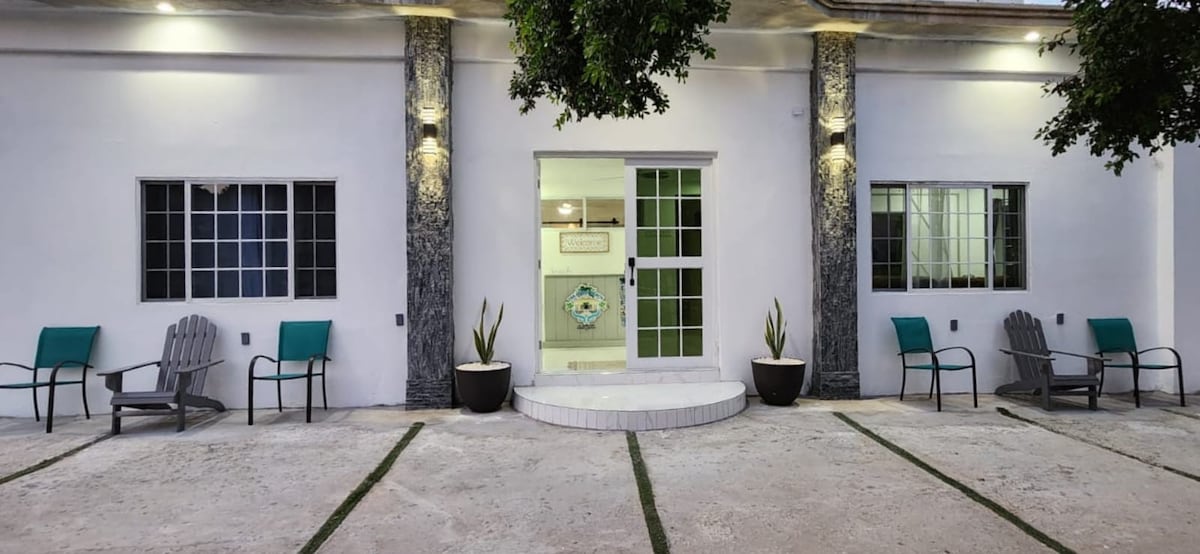 An exterior view showcases a modern building with a white façade and stone accents. Two large windows are framed on either side of the entrance, which features glass doors. Green chairs are positioned on a concrete area, with potted plants adding a touch of greenery.