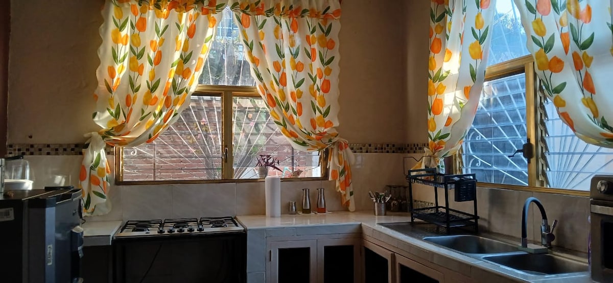A bright kitchen is presented, featuring a double basin sink and stove. Large windows are adorned with floral curtains, allowing natural light to fill the space. Countertops are equipped with essential utensils and a decorative metal rack for added organization.