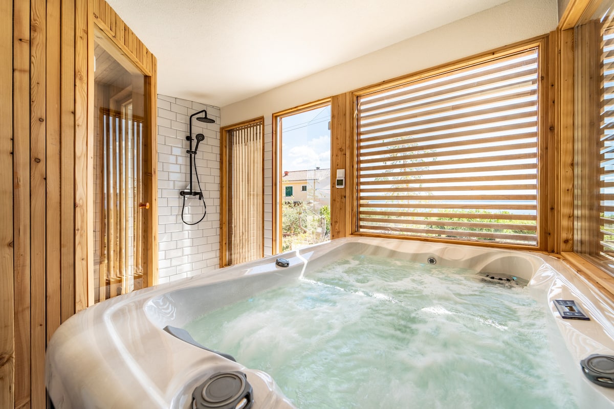 A spacious hot tub is positioned against wooden paneling, providing a calming atmosphere. Large windows allow natural light to fill the area, offering views of the garden. A shower is visible nearby, enhancing the wellness experience.