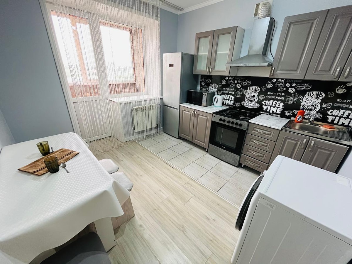 A modern kitchen is equipped with sleek gray cabinets and stainless-steel appliances, including a refrigerator and oven. A dining table with two place settings is positioned beside a window, allowing natural light to brighten the space. The wall features coffee-themed decor enhancing the kitchen's design.