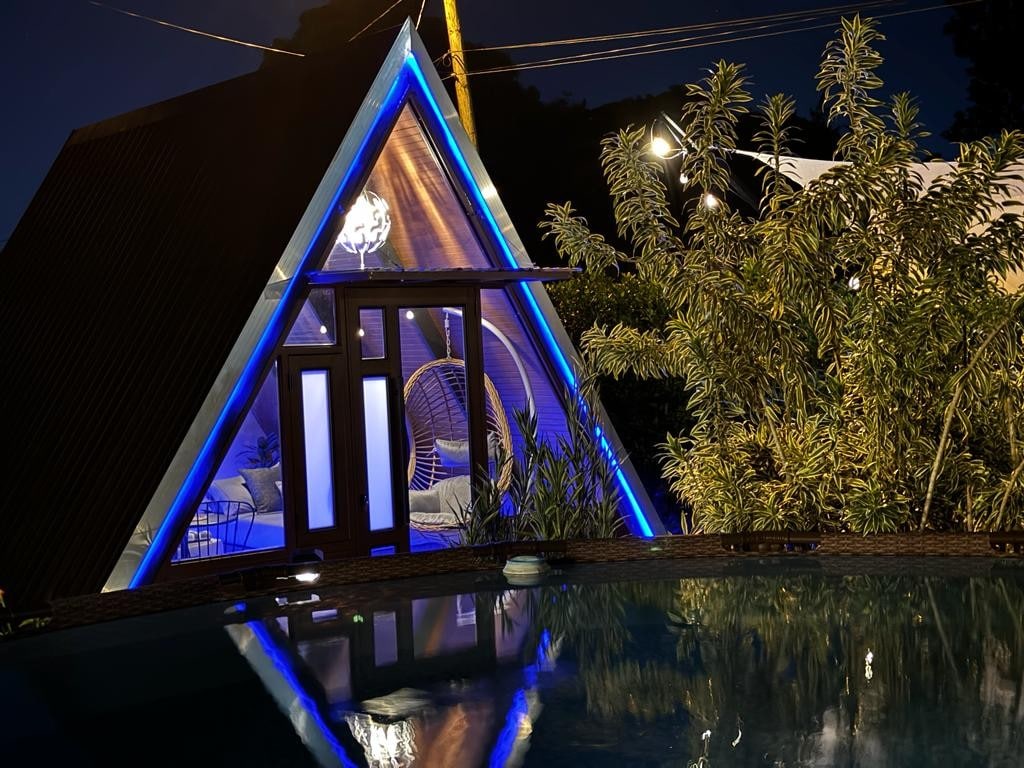An a-frame structure is illuminated by soft blue lighting, standing beside a tranquil pool reflecting its shape. The exterior features glass doors that open to a cozy living space, framed by lush greenery and fairy lights adding to the serene ambiance.