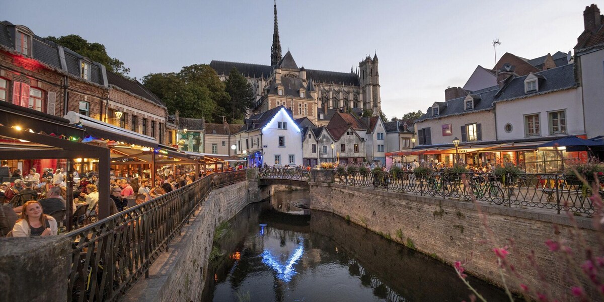 A vibrant scene captures a lively waterfront with restaurants lining the banks, illuminated by soft evening lights. Reflections dance on the water's surface, with a historical building visible in the background, showcasing architectural details. Trees are seen along the river, adding greenery to the atmosphere.