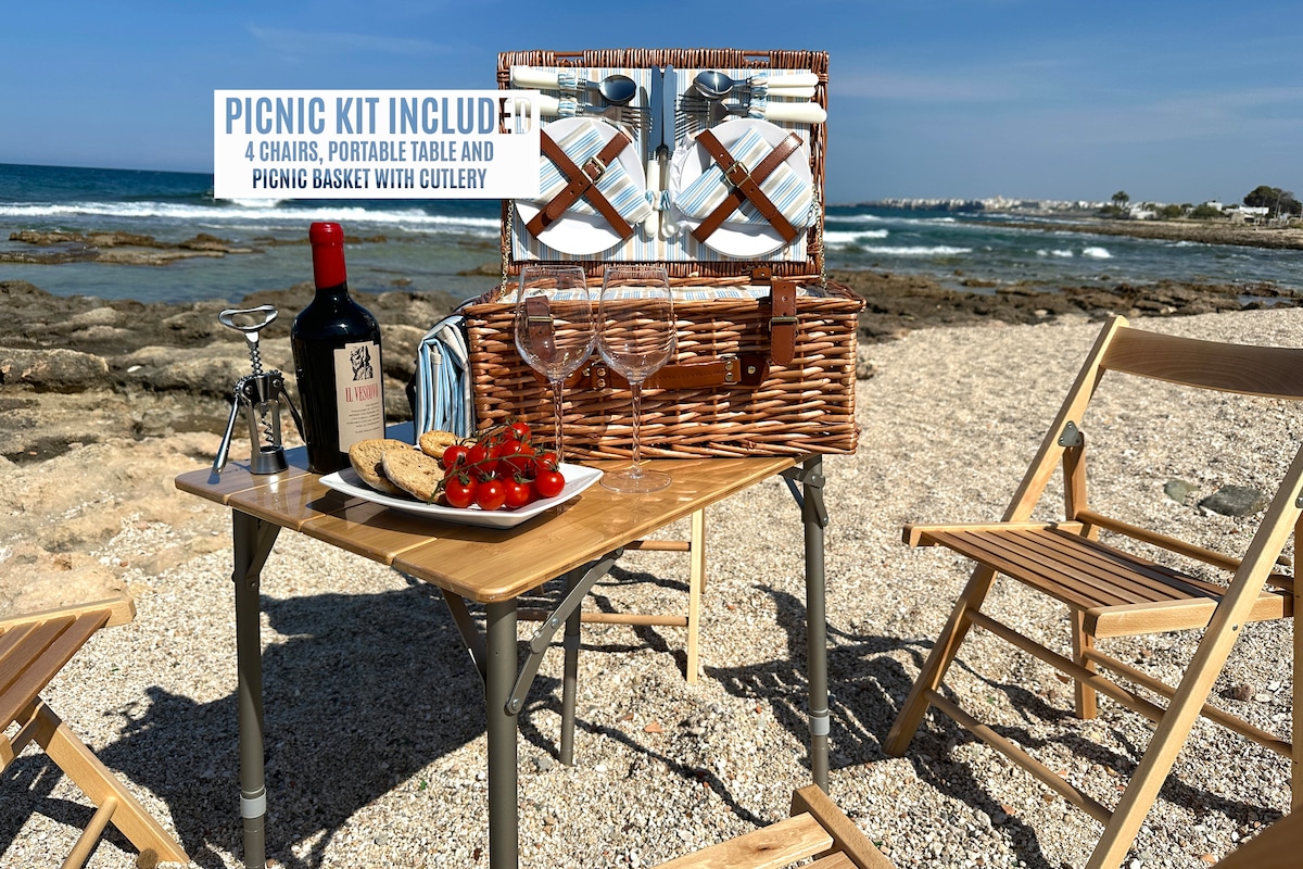 A portable picnic setup is presented on a sandy beach. The arrangement includes a small table with four folding chairs, a wicker picnic basket containing cutlery, two glasses, a bottle of wine, and a plate of snacks featuring tomatoes and cookies, with the sea visible in the background.