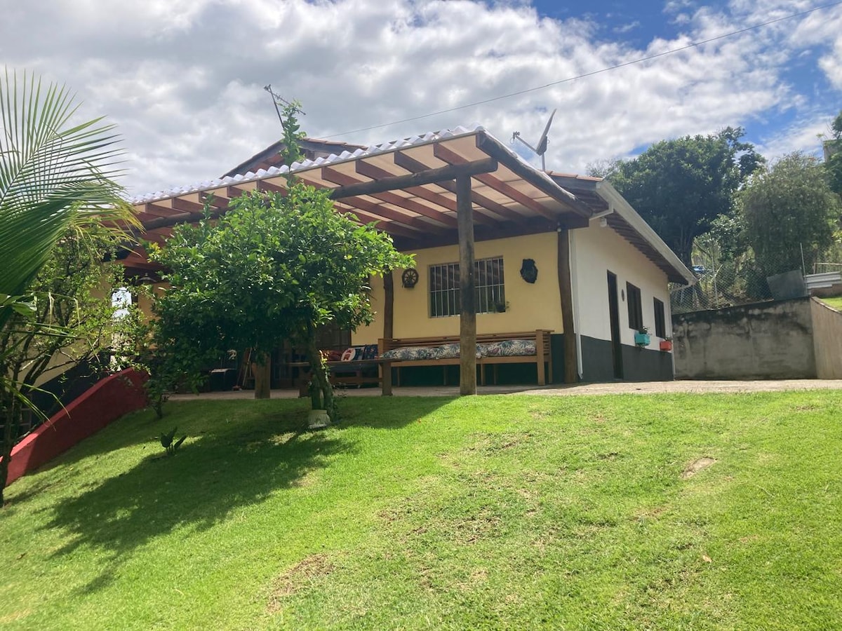 A spacious outdoor area features a covered porch with rustic wooden seating, surrounded by greenery. The vibrant lawn leads to a charming house with a white facade and dark trim, under a partially cloudy sky. The landscape includes trees and a gentle slope.