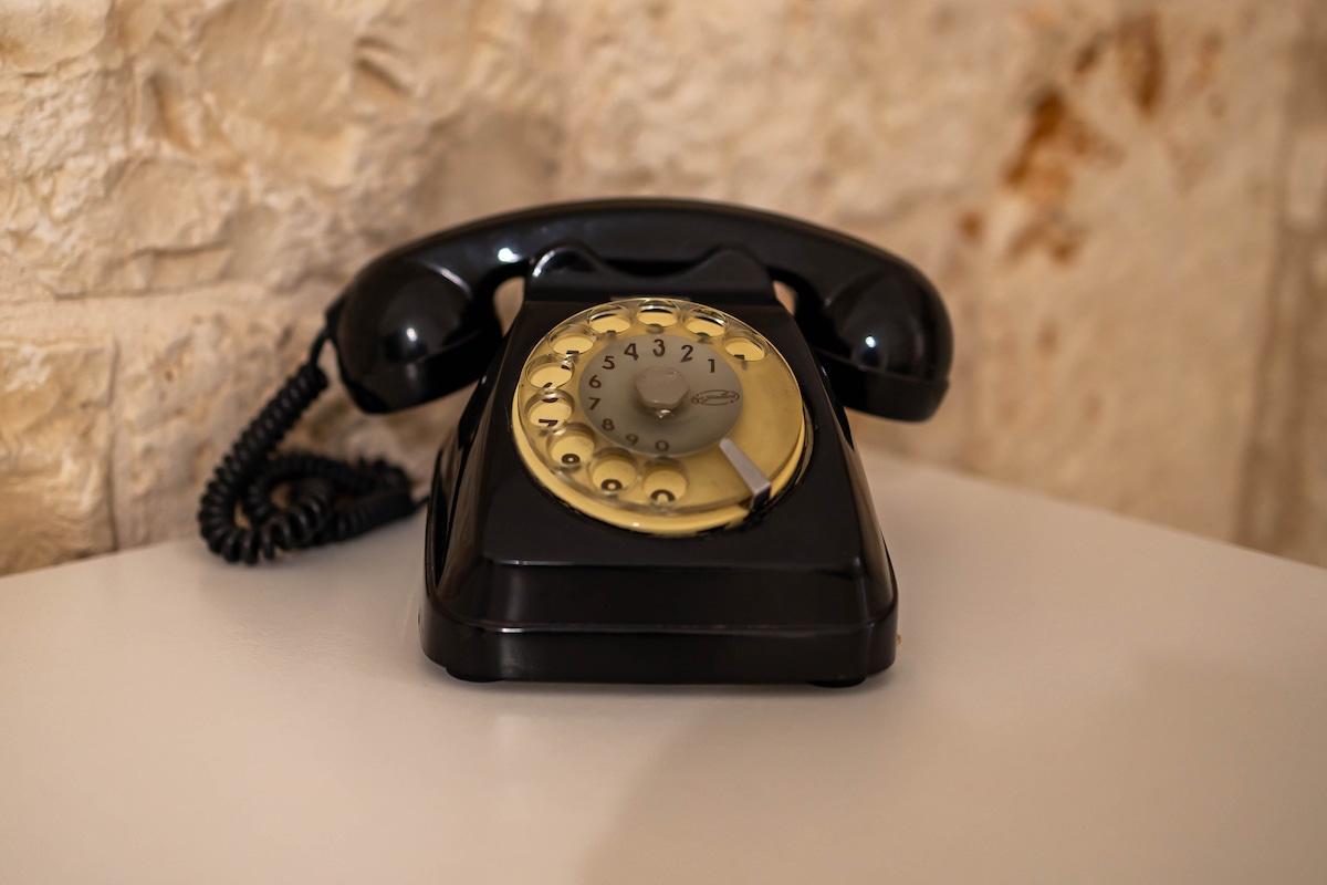 An elegant vintage rotary telephone is positioned on a sleek white surface. The telephone features a classic black body with a distinctive gold dial, showcasing the charm of retro design against a backdrop of natural stone walls.
