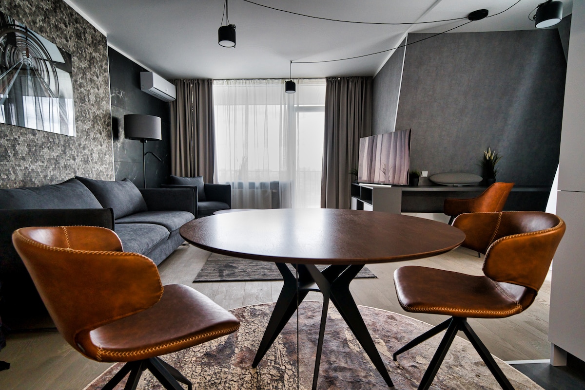 A modern living area is featured, showcasing a round wooden dining table surrounded by two brown leather chairs. A gray sofa is positioned next to a large window, draped with sheer curtains, allowing natural light to fill the space. A sleek TV console and decorative elements complete the room's design.