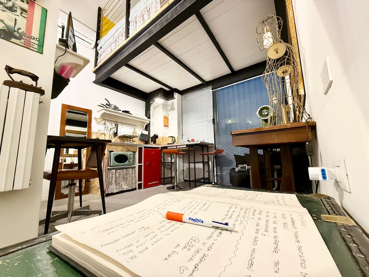 The interior space features a lofted sleeping area above a cozy living area. A collection of wooden furniture, including tables and chairs, is visible, while a kitchenette showcases red cabinetry. Natural light filters through the window, illuminating the room's creative atmosphere.