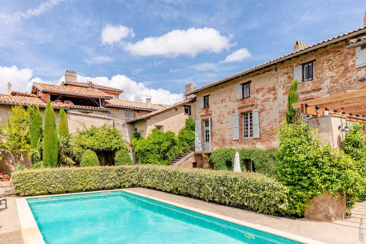 The image features a spacious outdoor area with a swimming pool surrounded by well-maintained greenery. The villa's rustic facade, comprised of textured stone and bricks, is visible, along with several trees and shrubs that enhance the tranquil setting. Bright blue skies hover above.