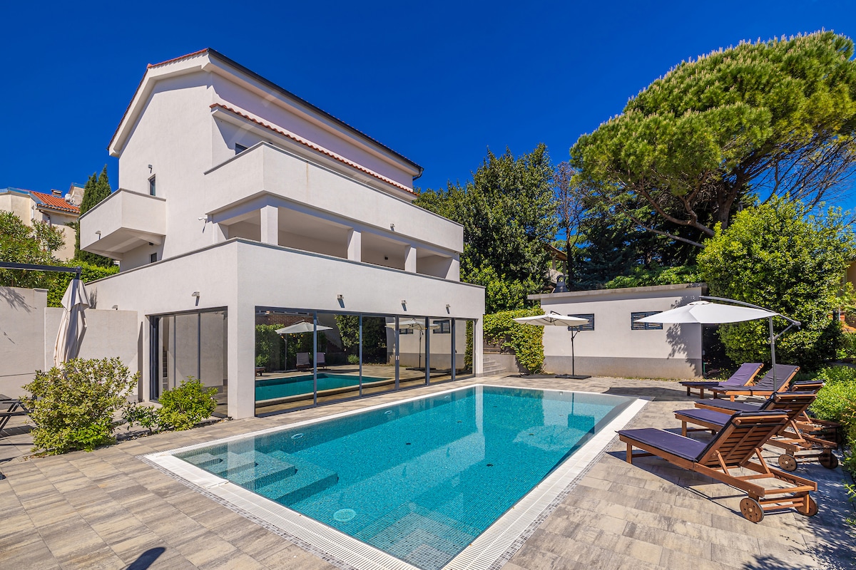 The exterior of the villa is highlighted by a spacious swimming pool surrounded by paved flooring. Sun loungers are positioned nearby, and large windows reflect the surrounding greenery, creating an inviting outdoor space. A shaded area with umbrellas provides additional comfort.
