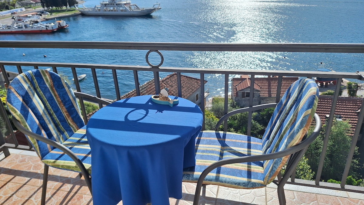 A private balcony provides a stunning view of the shimmering sea, accompanied by two striped chairs and a small round table set with a decorative item. The tranquil waters are visible alongside nearby houses and boats, enhancing the coastal atmosphere.