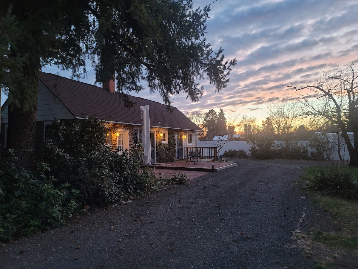 The exterior of the cottage features warm lighting as dusk approaches, with a spacious patio area visible. The gravel driveway leads toward the entrance, framed by trees and shrubs. A clear sky with soft clouds can be seen at sunset in the background.