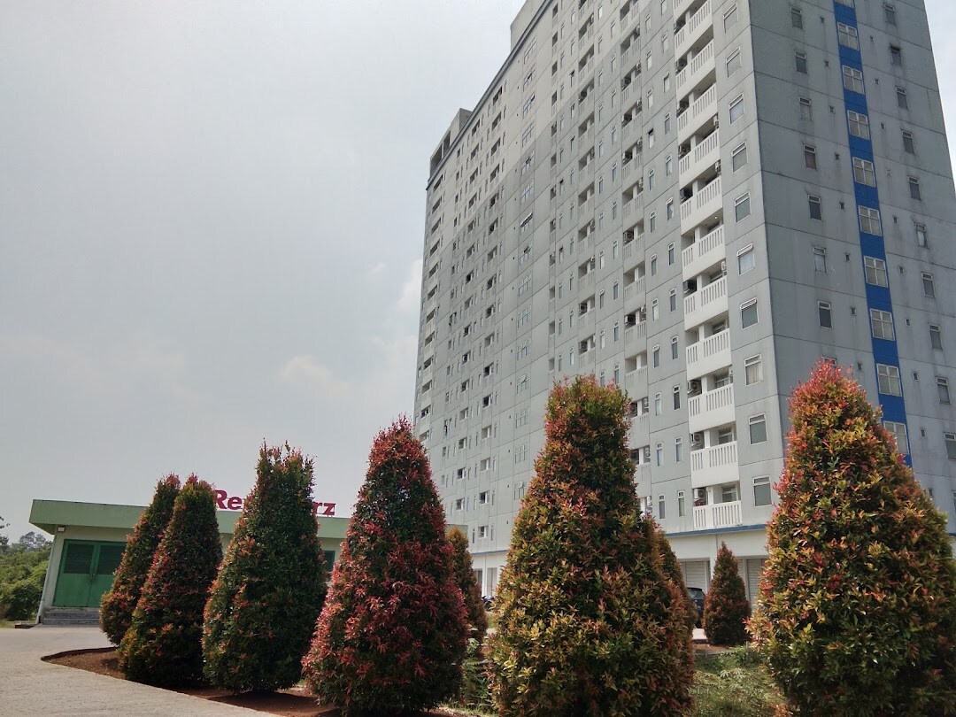 A modern multi-story building is surrounded by neatly trimmed bushes featuring vibrant red and green foliage. The exterior showcases a mix of light and dark colored panels, offering a contemporary look. The clear sky provides a bright backdrop to the overall scene.