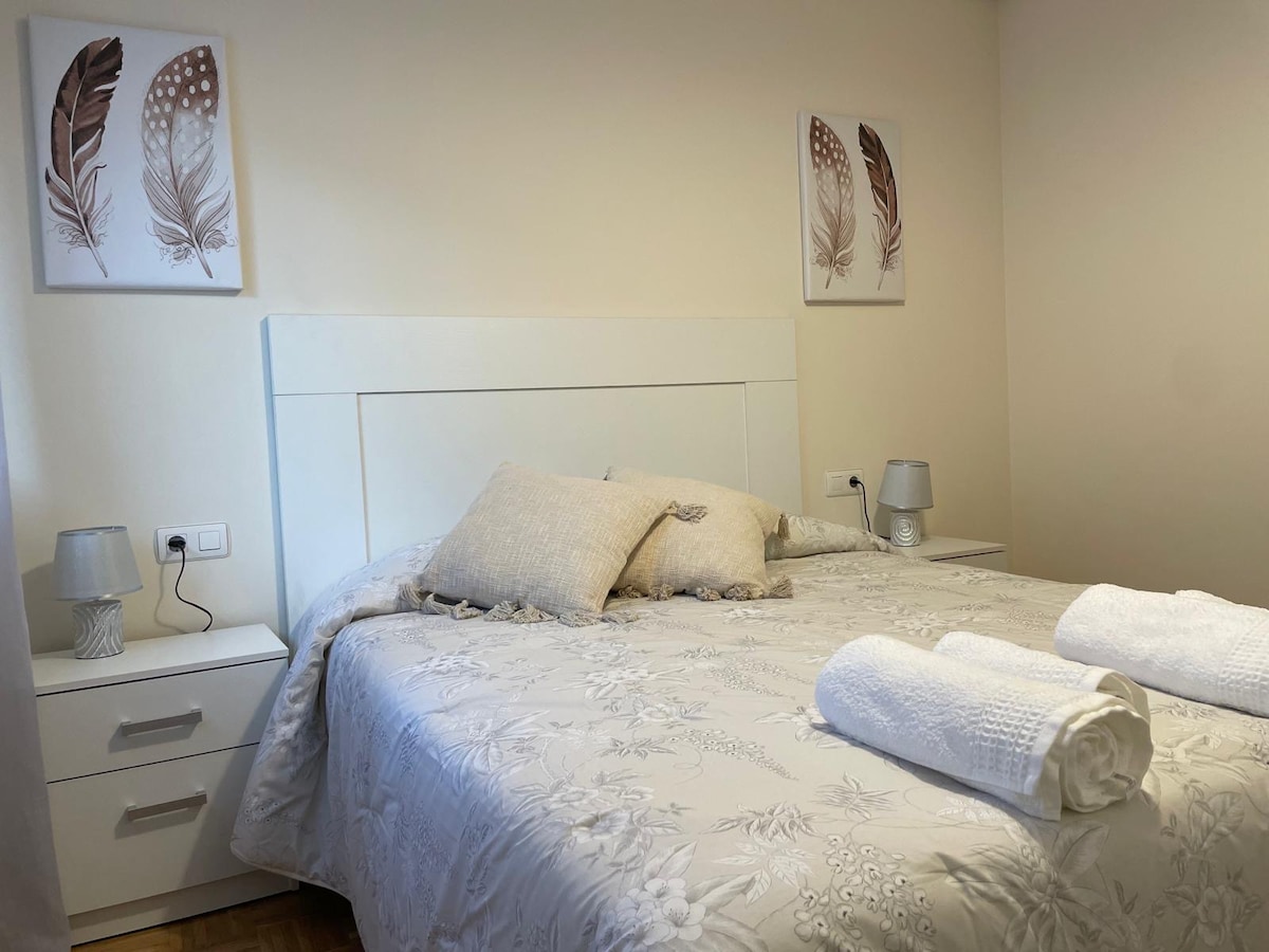 A well-appointed bedroom features a spacious bed centrally positioned, with a light-colored bedspread adorned with floral patterns. Nightstands with soft, ambient lighting stand on either side, complemented by decorative wall art depicting feathers.