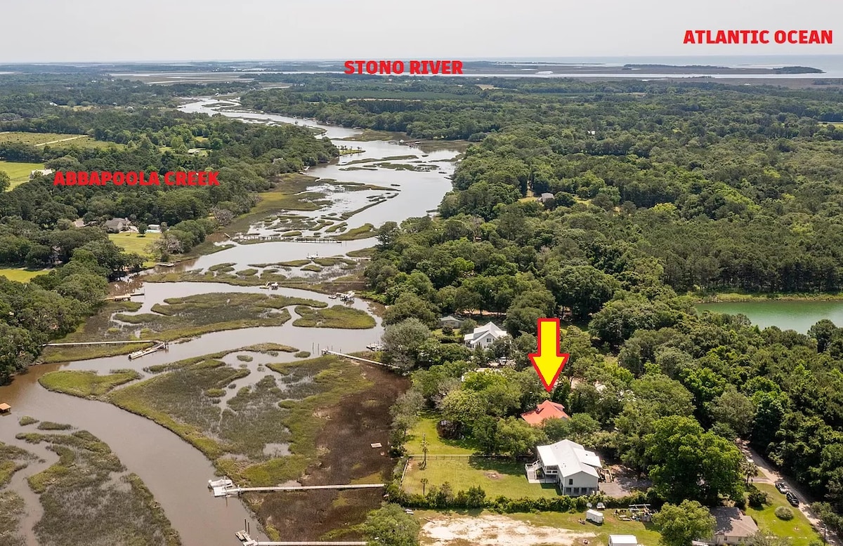 An aerial view captures the custom elevated cottage nestled on two acres, featuring expansive views of the tidal creek and Stono River. The Atlantic Ocean is visible in the distance, surrounded by lush greenery and waterways.