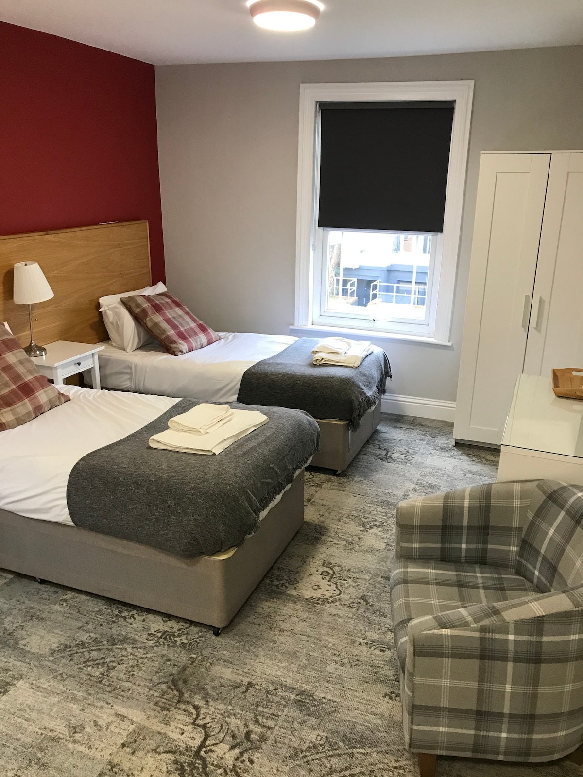 The room features two twin beds, each draped with a throw and neatly arranged towels. A small armchair is positioned near the window, which is covered with a roller blind. A wardrobe and a bedside lamp complete the decor, with a muted color palette enhancing the space.