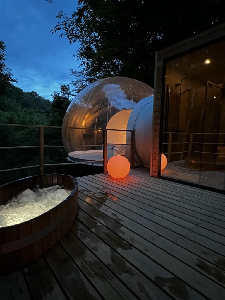 A wooden terrace showcases a unique transparent bubble structure, surrounded by lush greenery, under a twilight sky. A nordic bath filled with water and illuminated from within sits nearby. Soft glowing orbs provide subtle lighting, enhancing the tranquil atmosphere of the space.