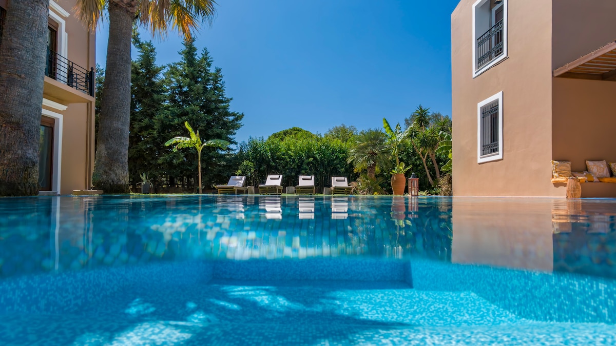 A serene swimming pool is captured, reflecting the clear blue sky. Three sun loungers are arranged by the poolside, surrounded by lush greenery. The villa's spacious exterior and a cozy seating area under a covered porch can be seen in the background.