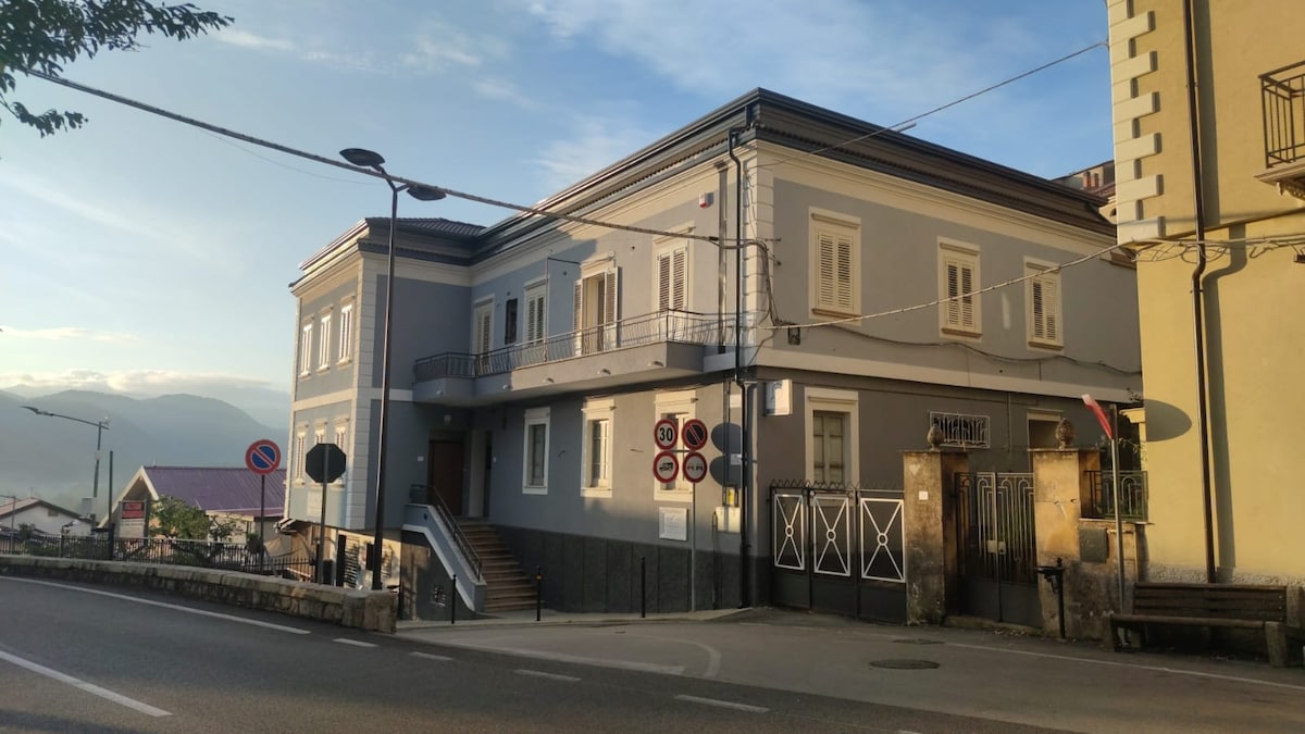 The building features a modern design with a light blue exterior and white window frames, located at the street corner. Multiple windows provide ample natural light, and a balcony enhances the facade. Surrounding trees and a roadway create a serene urban atmosphere.