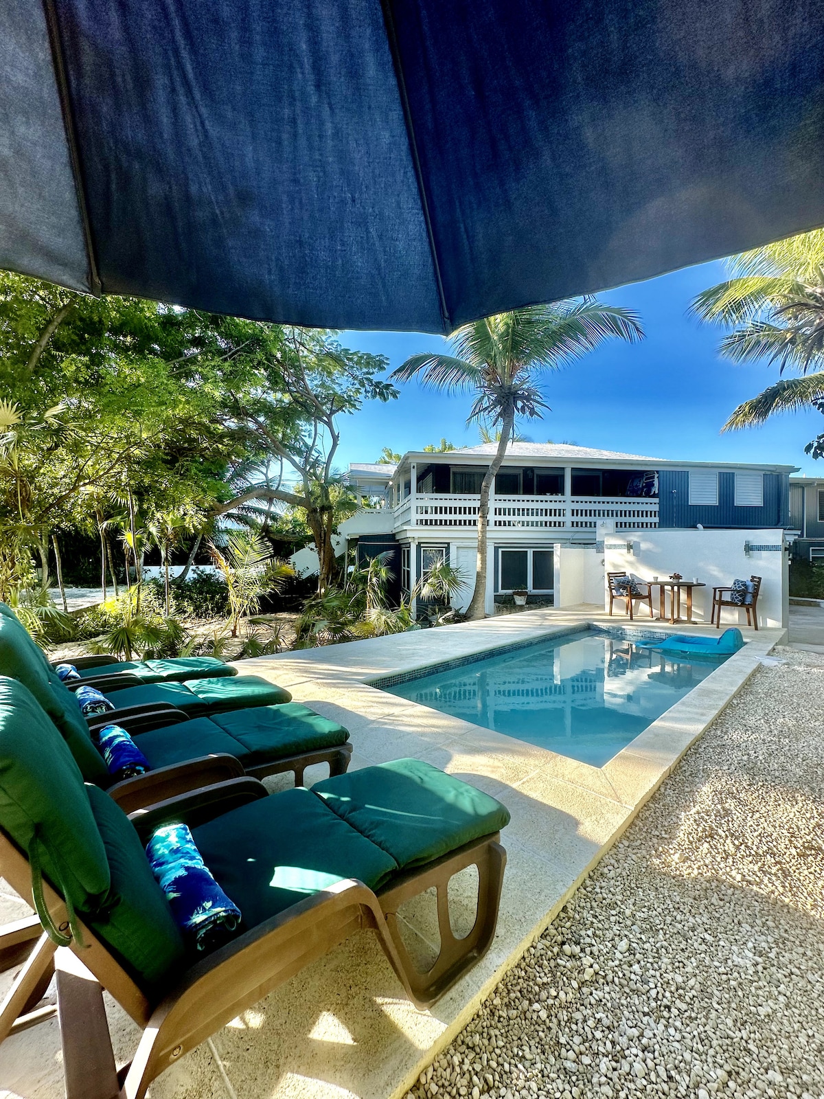A serene view showcases a swimming pool surrounded by lush greenery and lounge chairs under an umbrella. The back of a two-story villa is visible, featuring a spacious patio area with table and chairs, inviting relaxation in a tranquil setting.