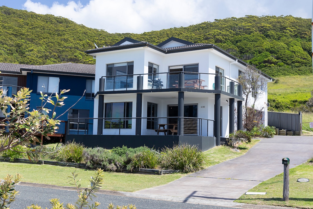 A modern two-story beach house is depicted, featuring a clean white exterior with large windows and balconies. Lush greenery surrounds the property, and a paved path leads to the entrance. The well-maintained lawn complements the serene landscape.