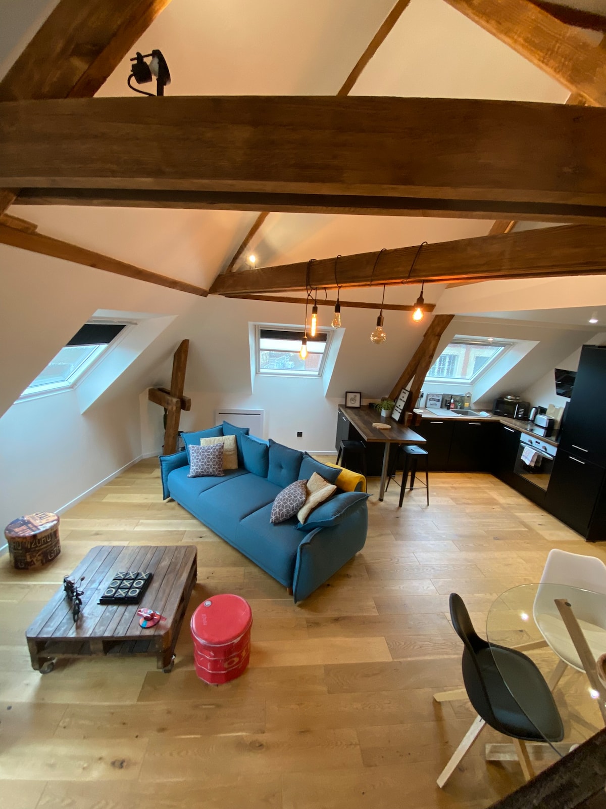 The loft space features a cozy blue sofa adorned with patterned cushions, set against a backdrop of wooden beams. A coffee table sits in front, with a casual dining area visible in the corner, complemented by modern kitchen appliances and large windows allowing natural light.
