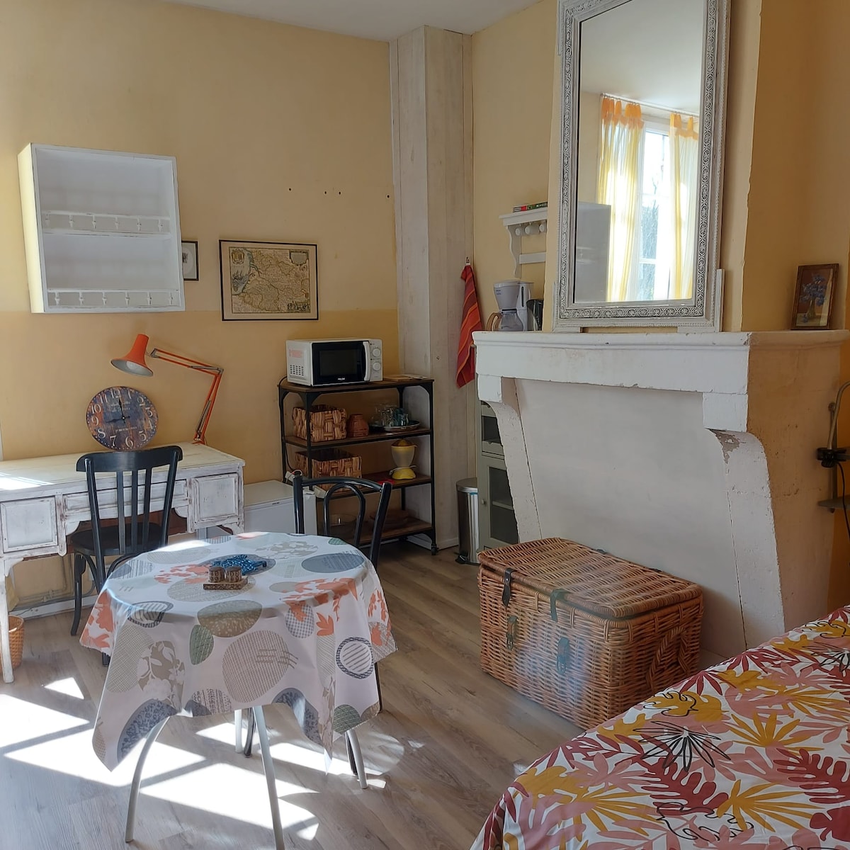 The room features a small dining table covered with a patterned tablecloth, accompanied by a black chair. A vintage-style mirror adorns the wall above a white fireplace. A microwave and other kitchen essentials are visible on a unit, alongside a window letting in natural light.