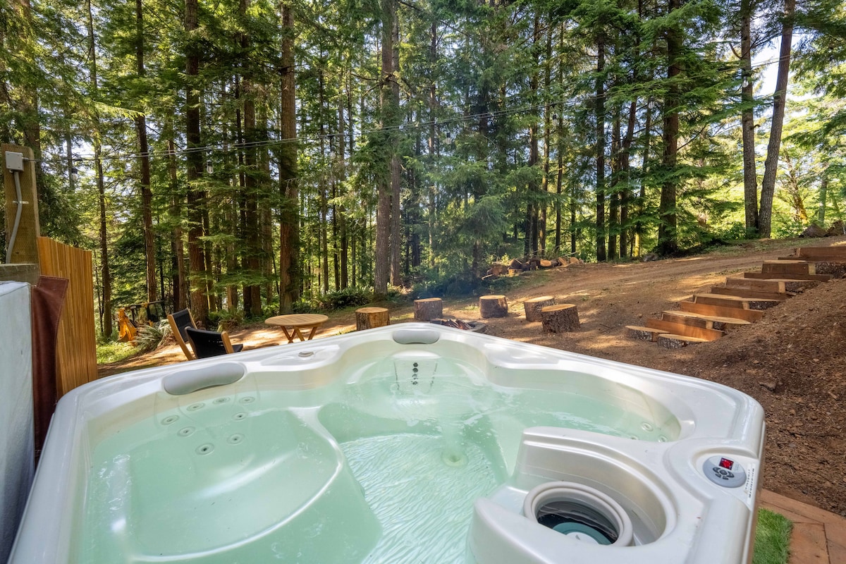 A private hot tub with clear water is surrounded by a forest of tall trees. In the background, a designated fire pit area with rustic seating is visible, offering an inviting space for relaxation in nature.