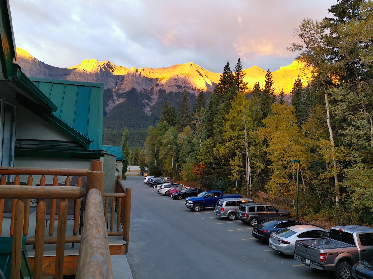 The image captures the stunning Rocky Mountains bathed in golden light during sunrise. A picturesque view of the forest is framed by towering pines, with a parking area visible in the foreground showcasing various vehicles.