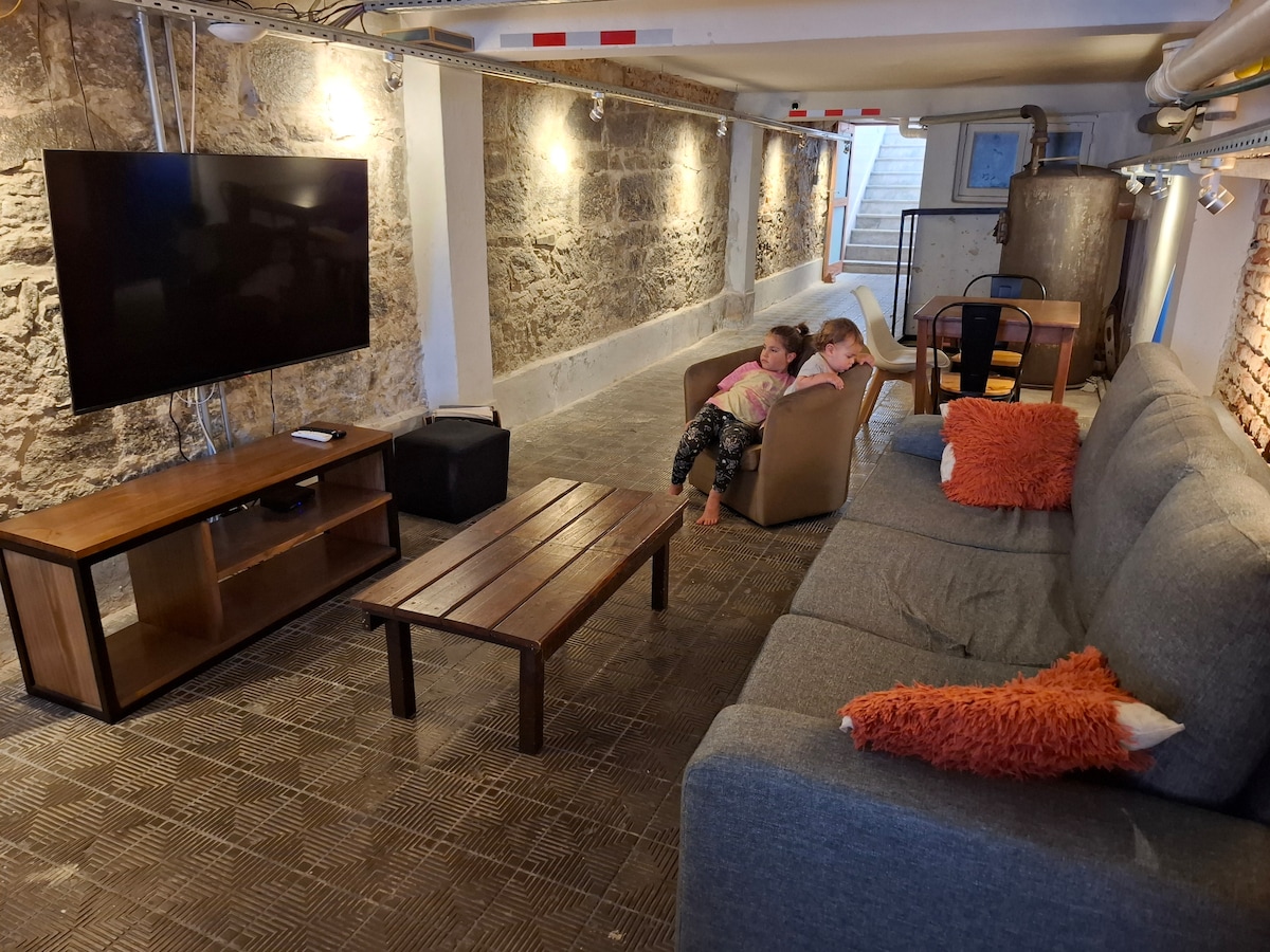 A lower level game room is featured, showcasing a large flat-screen TV mounted on the wall. A gray sectional couch and two additional chairs are arranged around a wooden coffee table, enhancing the space's comfort. Exposed stone walls add character, while bright lighting illuminates the area.
