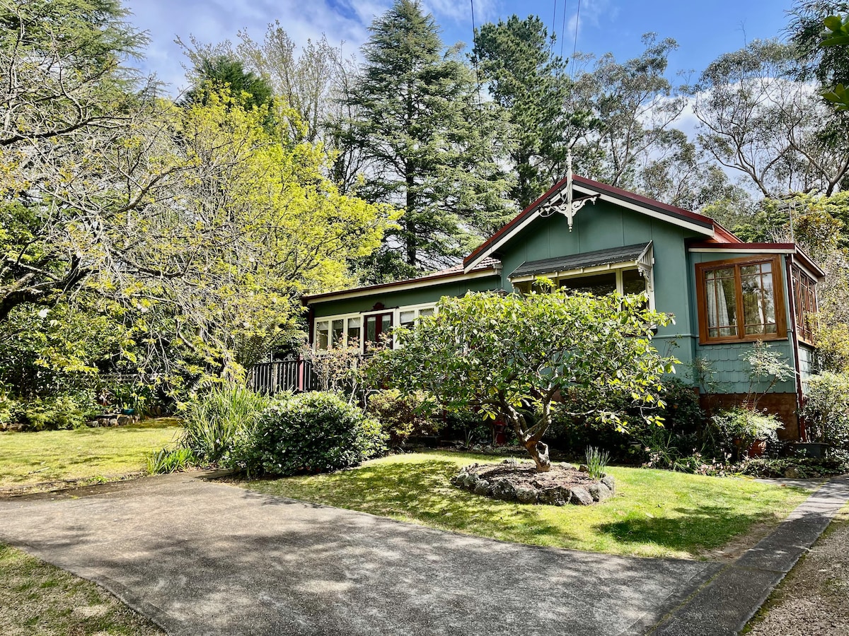 The exterior of Rose Cottage is framed by lush greenery and vibrant trees, showcasing a charming single-story structure. A paved pathway leads to the entrance, with a well-maintained lawn and small landscaped areas complementing the peaceful surroundings.