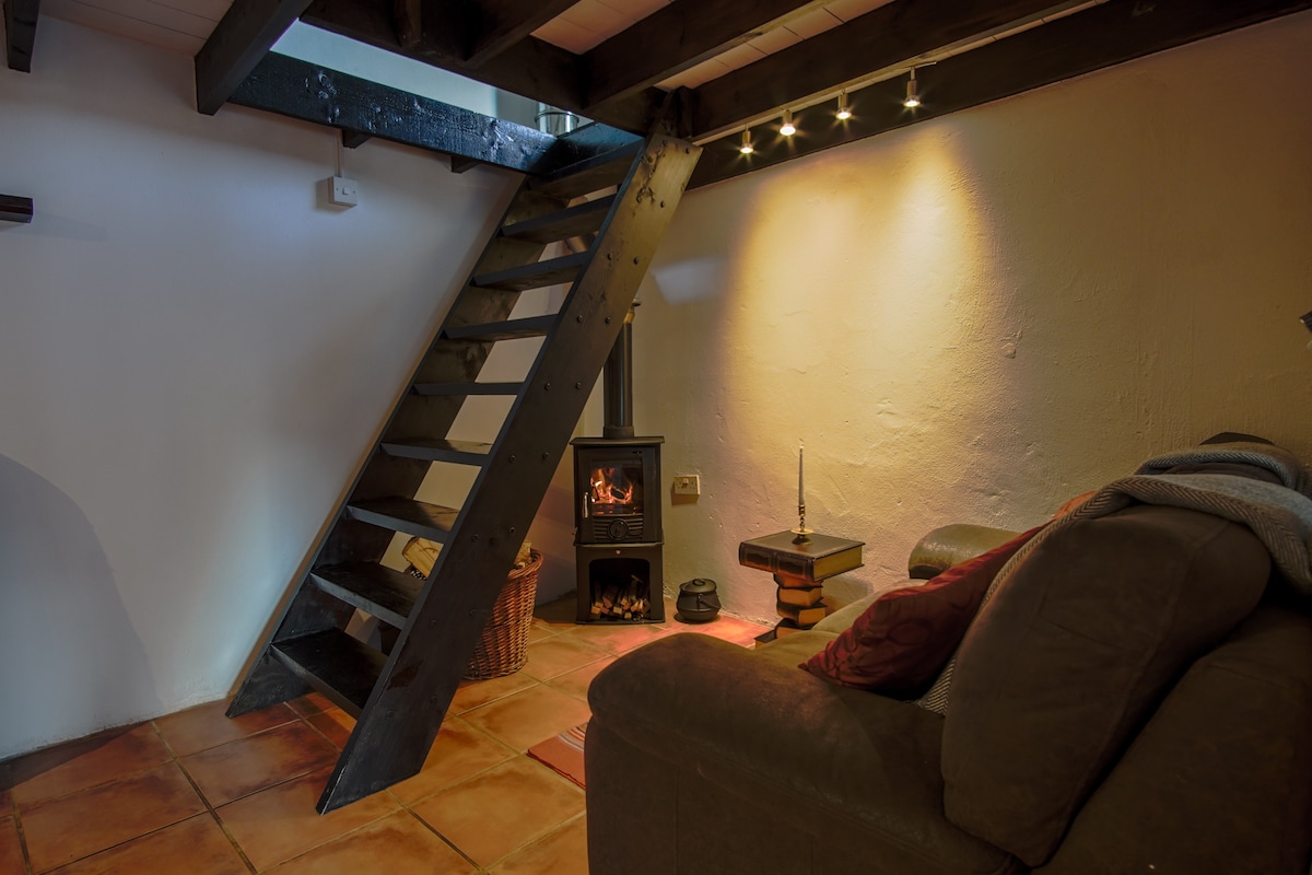 A wooden staircase leads to the upper floor, with a cozy living area featuring a brown sofa. A wood-burning stove adds warmth to the space, complemented by soft lighting and a textured wall, creating a comfortable and inviting environment.
