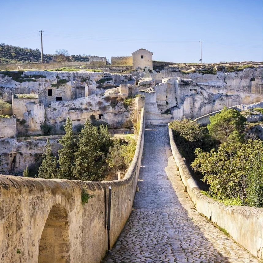 A scenic view captures the ancient landscape of Gravina in Puglia, featuring a stone bridge leading toward a traditional building atop a rocky outcrop. The terrain is marked by cliffside caves and lush greenery, under a clear blue sky.