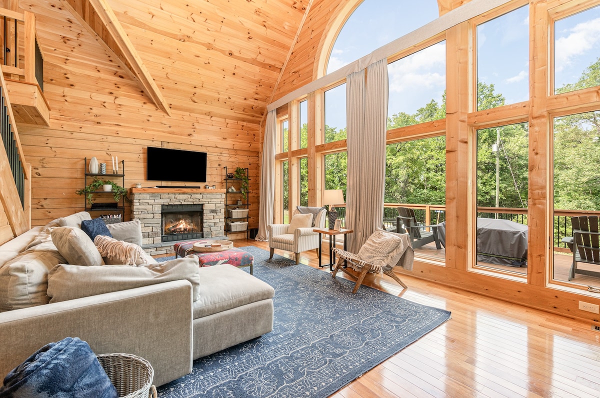 A spacious living area features wooden walls and high ceilings, with large windows allowing natural light. A comfortable sectional sofa is positioned near a stylish fireplace, while a cozy armchair and a side table enhance the inviting space. Outdoor seating is visible through the glass doors.