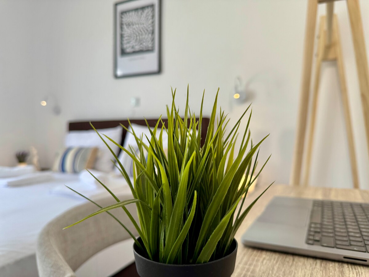 A small pot of lush green grass is foregrounded on a light wooden table. In the background, a cozy bedroom with a spacious bed is visible, dressed in fresh linens. A framed artwork hangs on the wall, and soft lighting is provided by wall-mounted lamps.