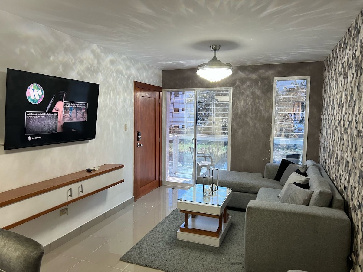 A living area features a gray sectional sofa arranged around a glass-topped coffee table. A large television mounted on the wall displays content, while natural light enters through the windows. A door leads outside, and the space is accented by a contemporary ceiling light.