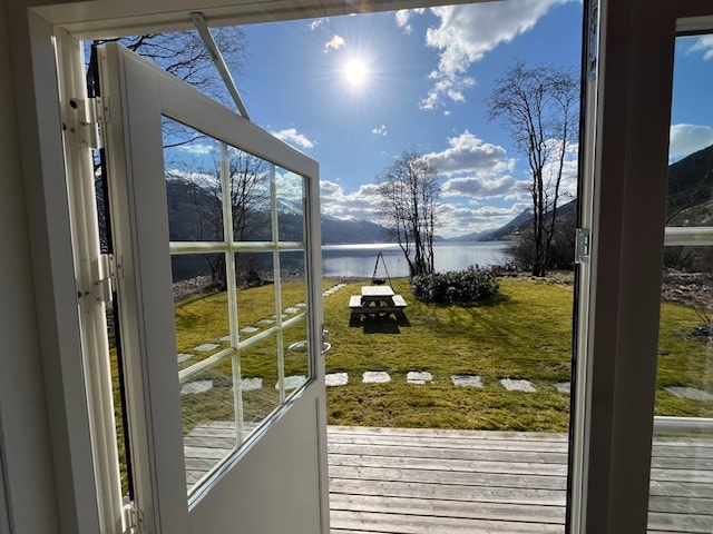 An open door reveals a scenic view of the lake and surrounding landscape. A picnic table is visible on the lush grass, with pathways leading towards the water. The bright sky, illuminated by sunlight, enhances the tranquil outdoor setting.