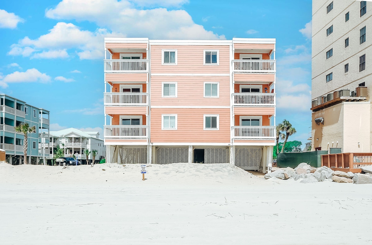 A three-story coral-colored building stands directly on the beach, featuring balconies on each level. The sand is visible in the foreground, and a clear blue sky enhances the beachside setting. Nearby structures are also visible, contributing to the coastal atmosphere.