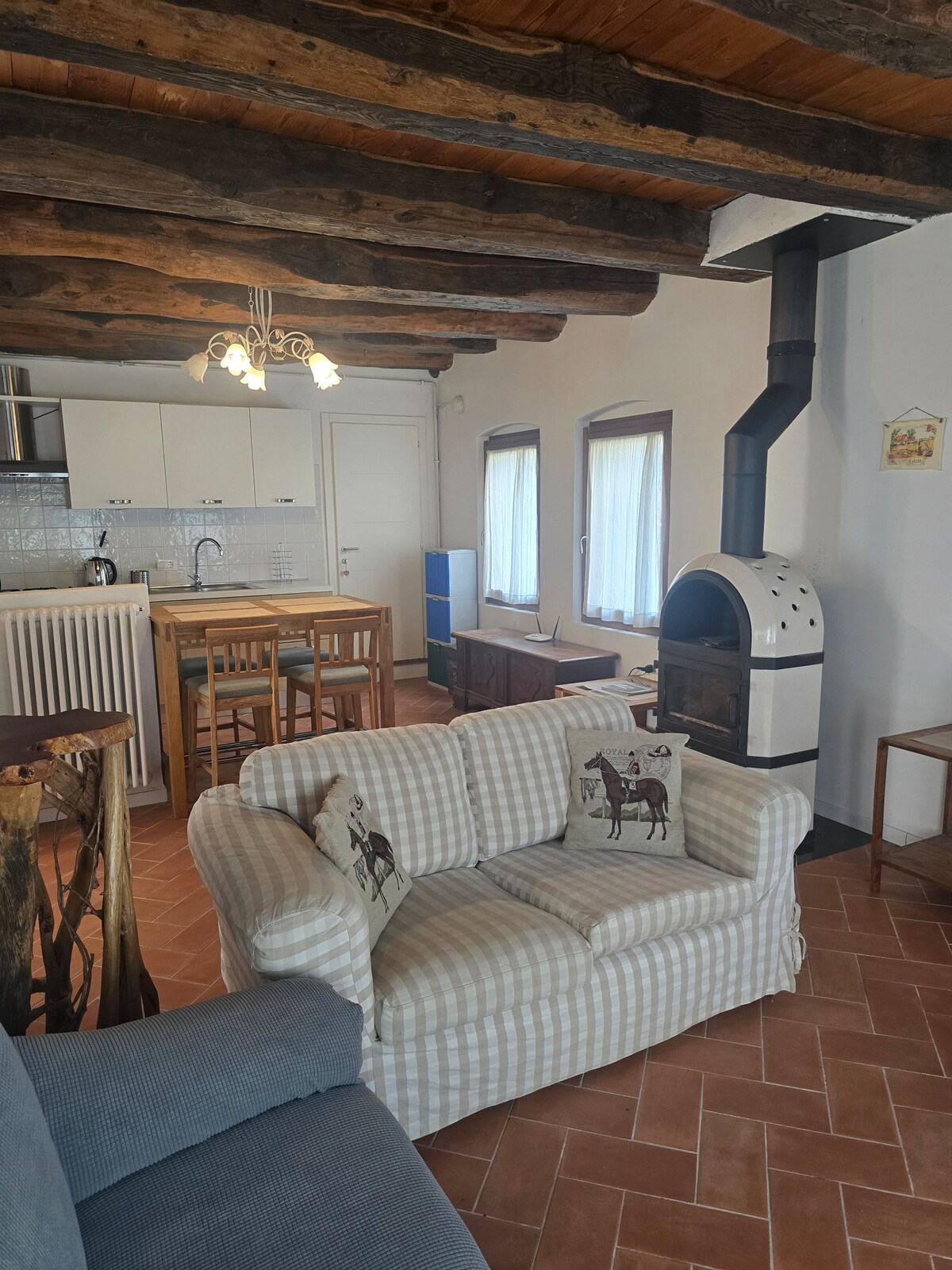 A warm living area features exposed wooden beams overhead and terracotta tile flooring. A patterned couch with decorative pillows and an armchair provide seating. A dining table with chairs is positioned near the kitchen, and a wood-burning stove stands against the wall beside the windows.
