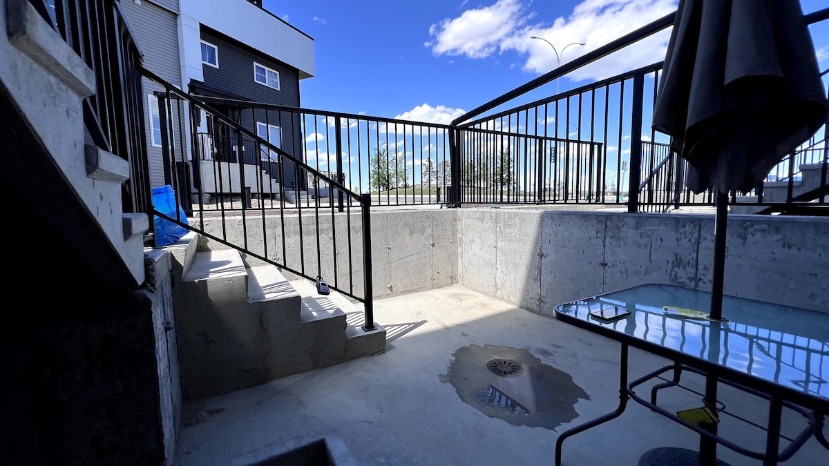 A private outdoor area is depicted, featuring a concrete surface surrounded by a black railing. Steps lead down to the space, which has a small drainage area. A glass table and umbrella are visible, providing a practical setting for outdoor relaxation.