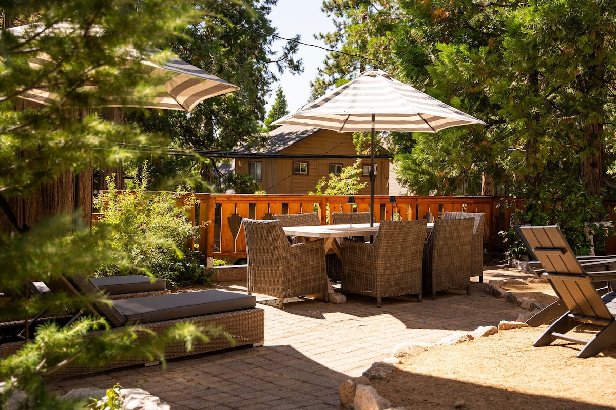 A spacious outdoor area is furnished with a wooden dining table surrounded by wicker chairs, shaded by two large umbrellas. Lounge chairs are positioned nearby on a stone walkway, bordered by greenery and pine trees, creating a serene atmosphere.