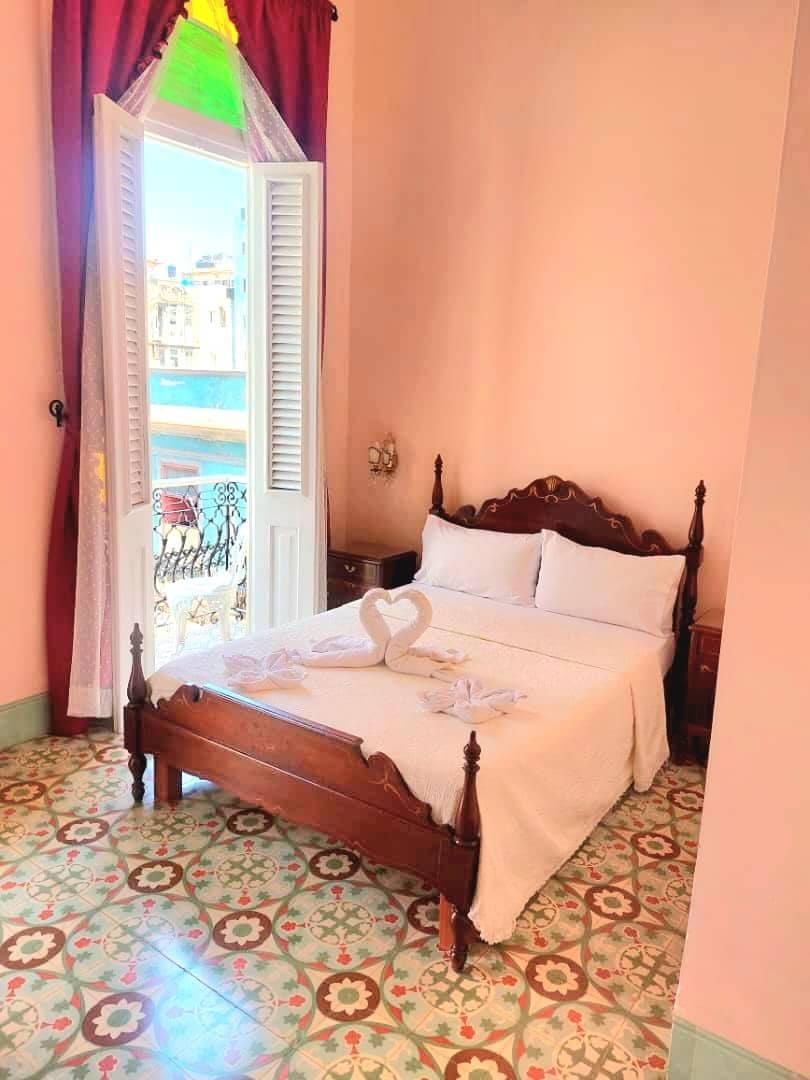 A cozy bedroom features a wooden bed with a light blanket arranged decoratively. Large windows with shutters open to a scenic view, allowing natural light to fill the space. Colorful floor tiles enhance the room’s charm, complemented by soft pink wall tones and rich curtains.