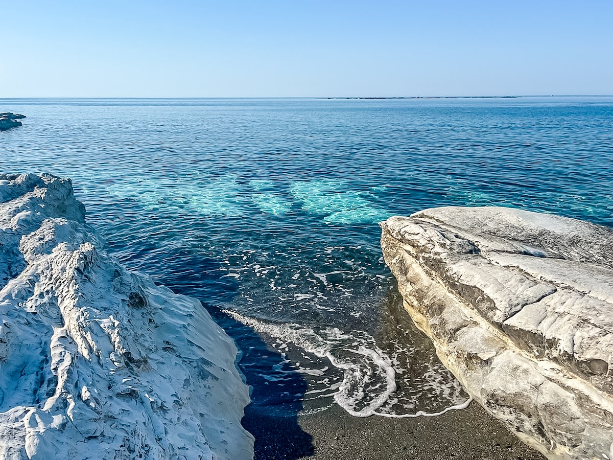 A serene coastal scene depicts calm waters reflecting clear blue skies. White rocks frame the shoreline, with gentle waves lapping against the smooth surfaces. The horizon stretches into the distance, meeting the sea where the sky and water merge seamlessly.