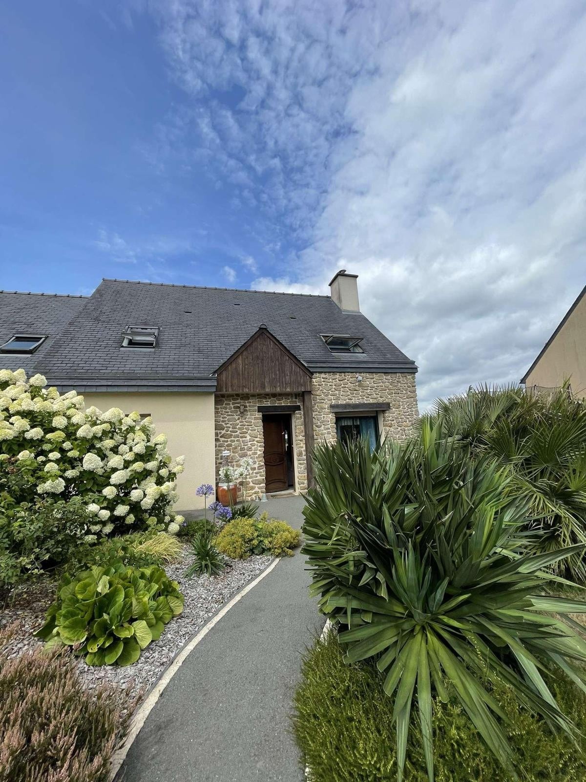 An exterior view of a charming house is surrounded by a landscaped garden featuring a variety of greenery and blooming flowers. A curved pathway leads to the entrance, framed by lush plants and shrubs. The sky above is partly cloudy, indicating a pleasant atmosphere.