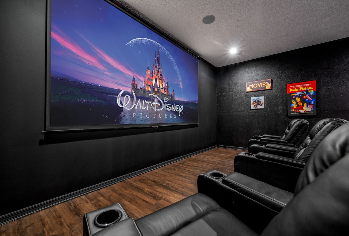 A private movie theater features a 150-inch screen displaying a Disney image. Black walls and hardwood flooring create a modern atmosphere. Comfortable reclining seats with cup holders are arranged for a cinematic experience, ensuring a cozy and immersive viewing environment.